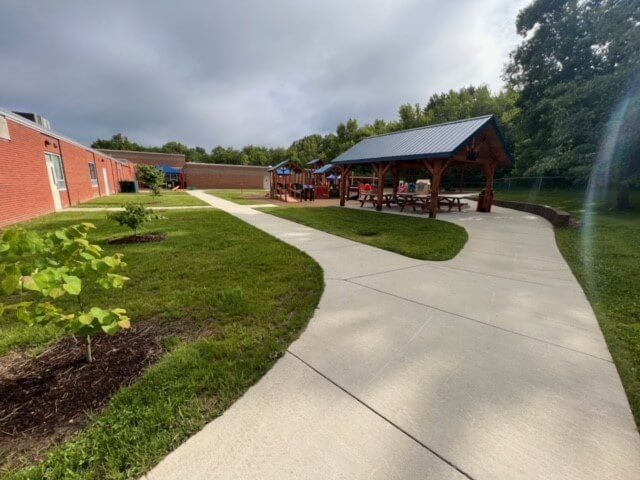 A concrete path leads past a red brick building and a wooden shelter with picnic tables. Playground equipment is visible in the background.