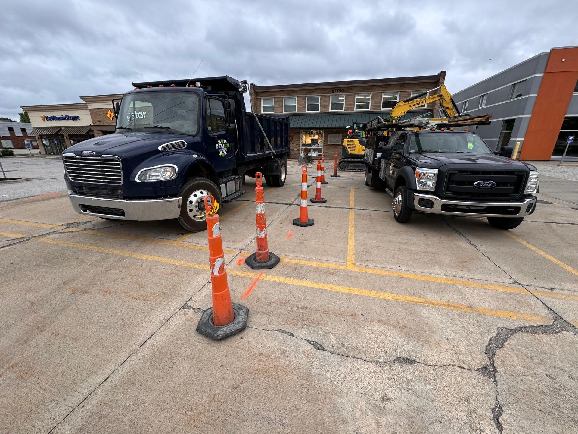 Two work trucks and construction equipment in a parking lot, marked by orange cones. A two-story brick building is in the background.