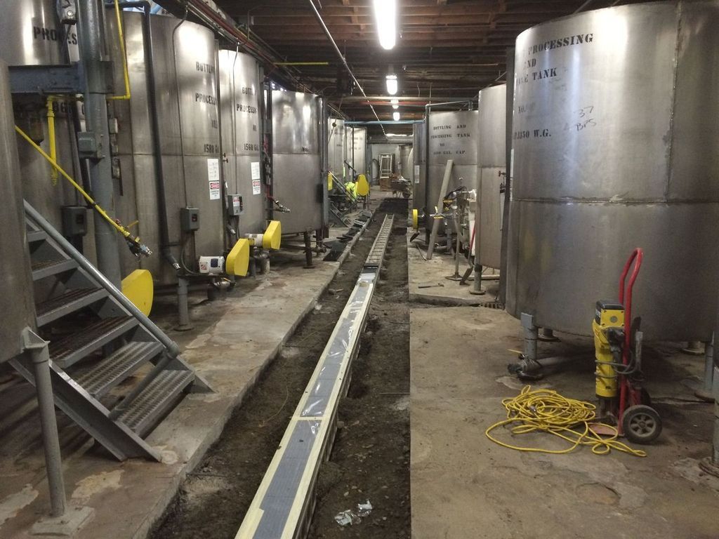 Inside a winery with stainless steel tanks, a central drainage channel, and industrial equipment. The setting appears dimly lit with yellow accents.