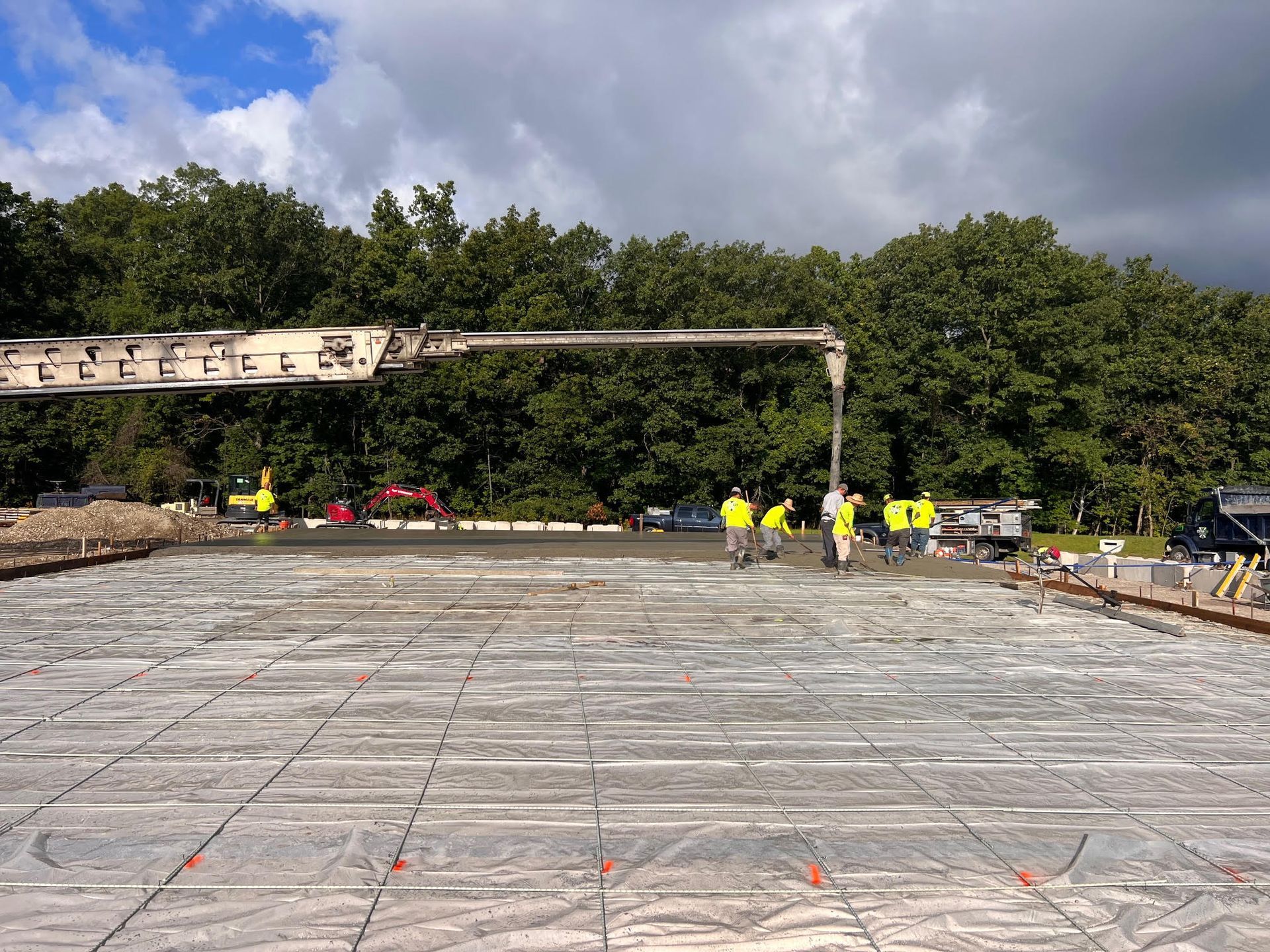 Construction workers pouring concrete for a foundation. A large pump arm extends over the work area, with trees in the background.