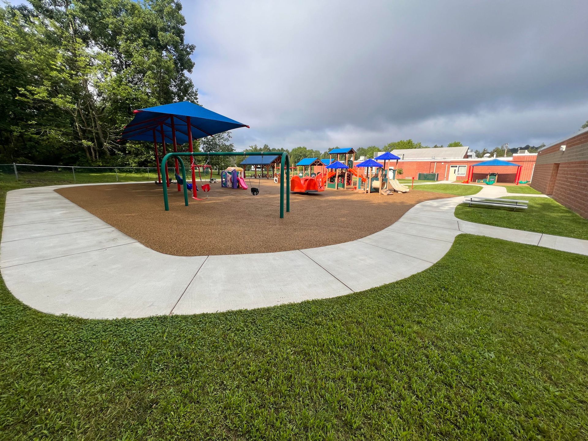Playground with swings and slides; brown safety surface, a paved path, green grass, and a cloudy sky.