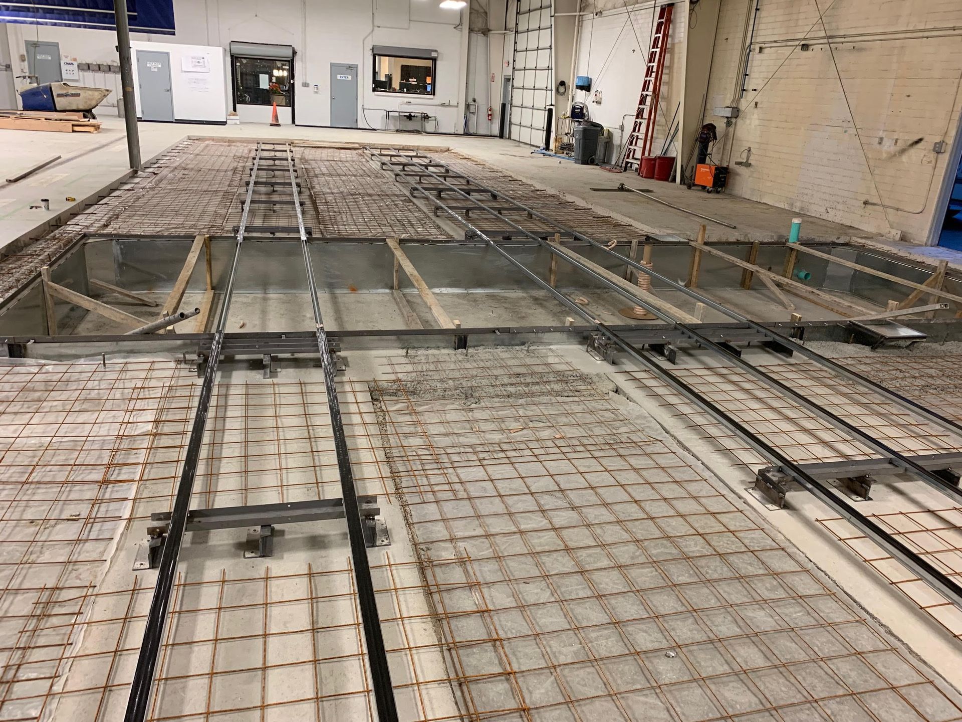 Construction site with a metal framework and rebar grid, possibly for a concrete floor. Industrial setting with white walls and equipment visible.