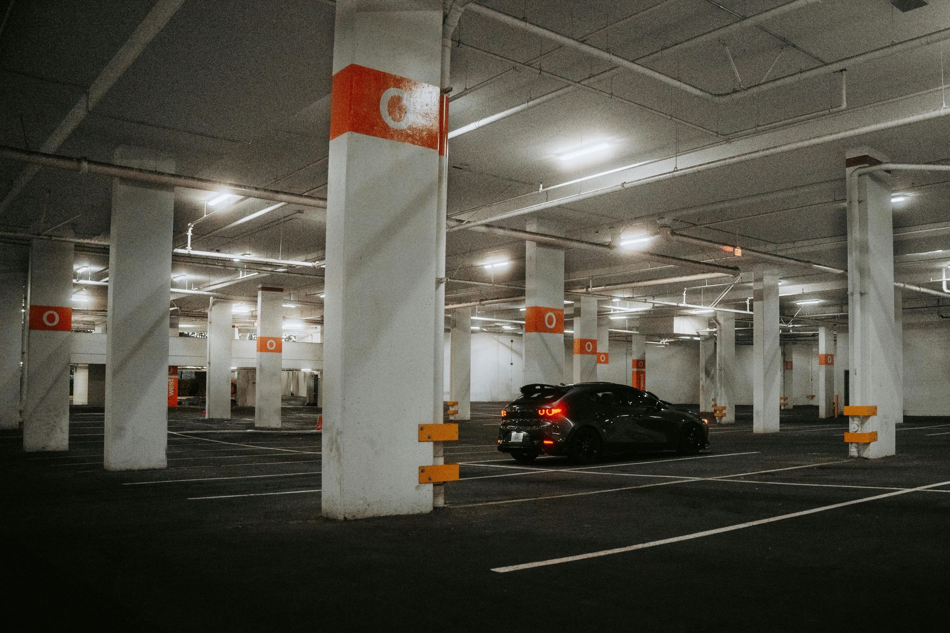 Black car in a mostly empty, brightly lit parking garage; columns with orange markings.