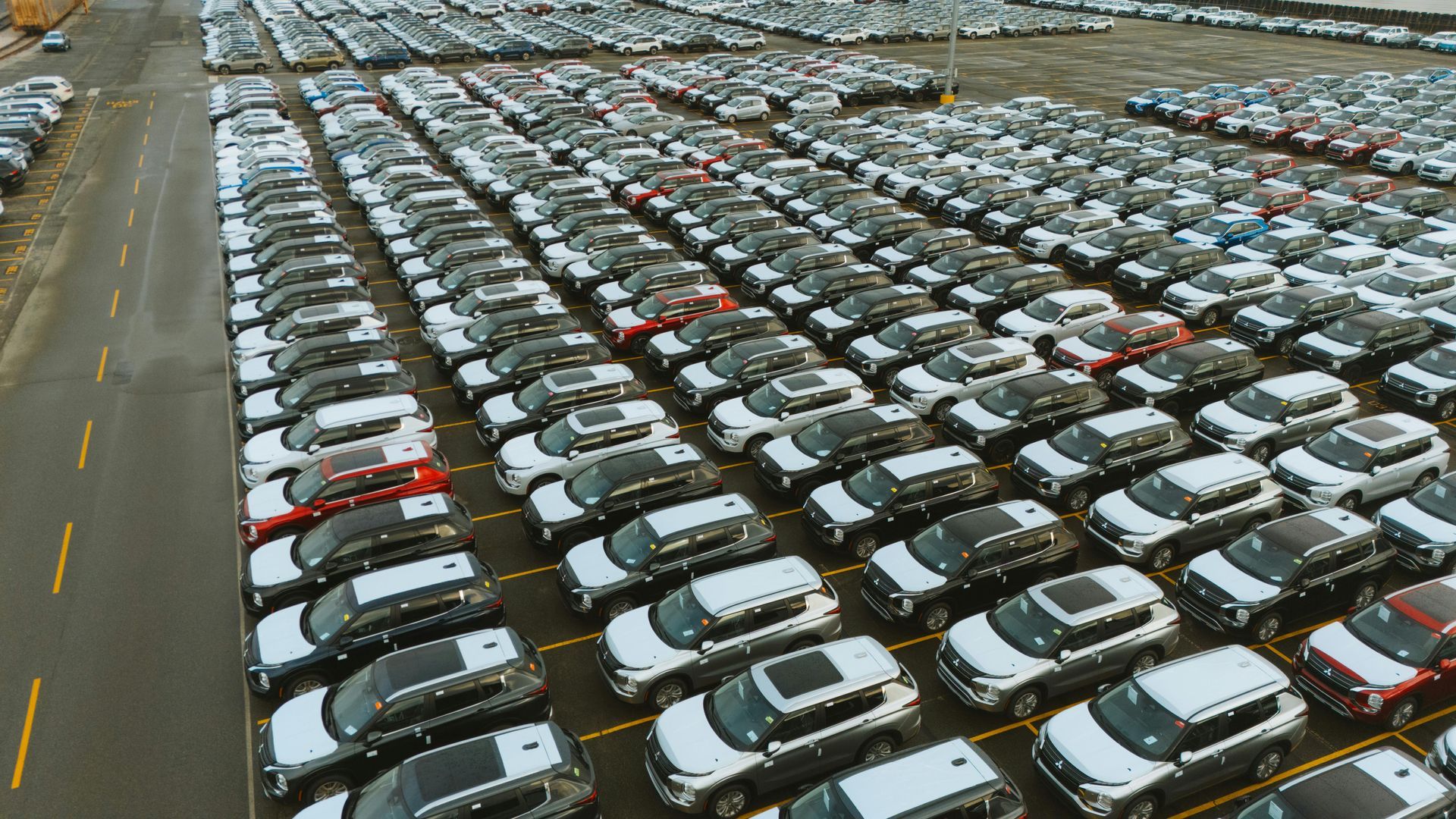 Rows of new cars parked in a large outdoor lot; mostly white, gray, and red vehicles.