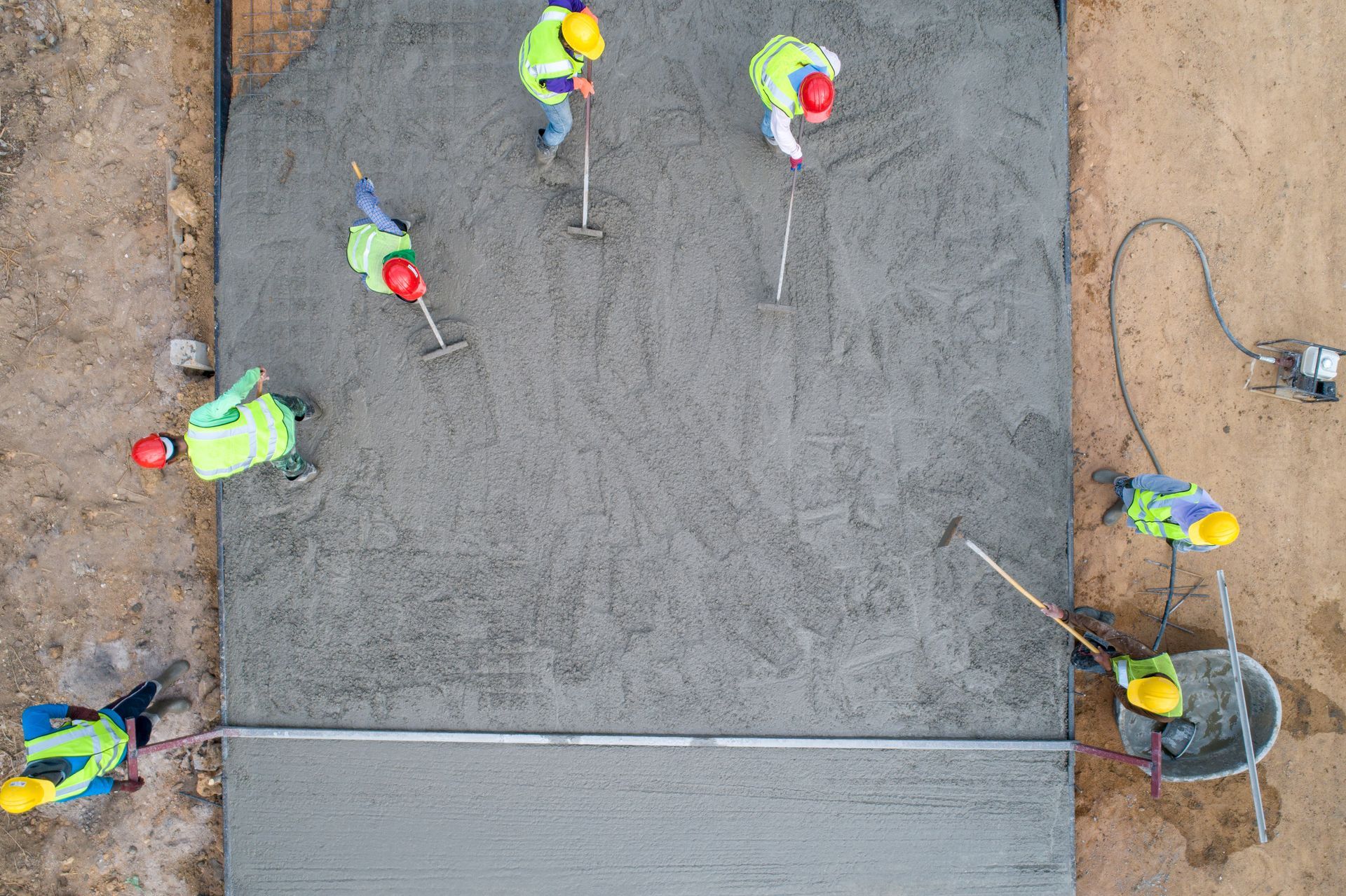 Construction workers smoothing wet concrete on a road, wearing safety vests and helmets, working in an outdoor setting.