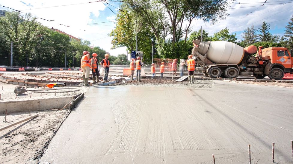 Construction workers pouring concrete on a road, with a cement truck present. The workers wear orange vests and hard hats.