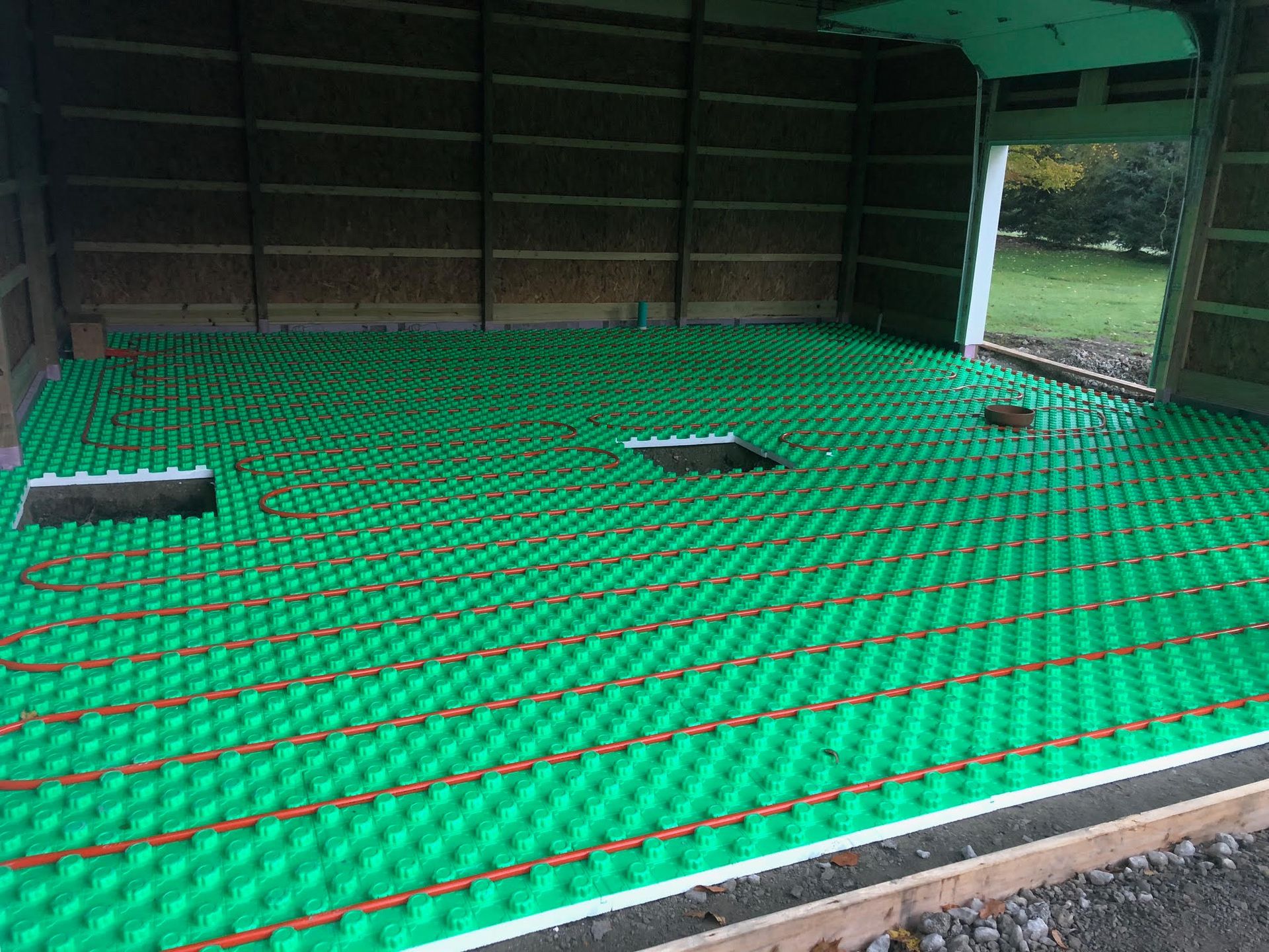 Green flooring with copper pipes, ready for concrete, inside a wood-framed building. Open doorway leads to a grassy outdoor space.