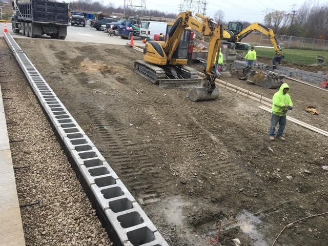 Construction site with heavy machinery and workers. Excavators move dirt, a cinder block wall is being built next to gravel.