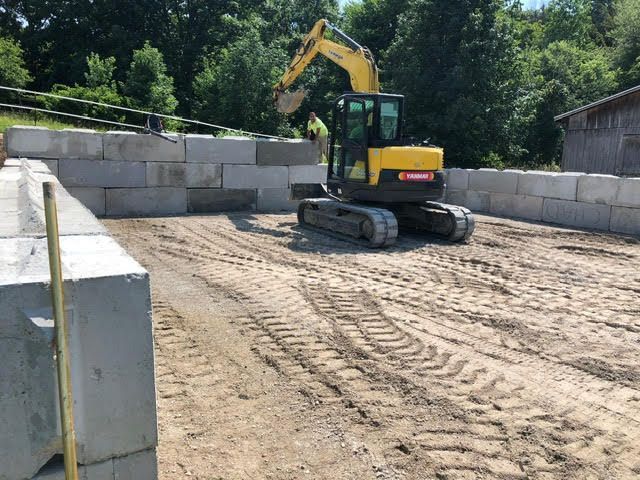 A yellow excavator positioned in a sandy area between concrete retaining walls. It appears to be doing construction work.
