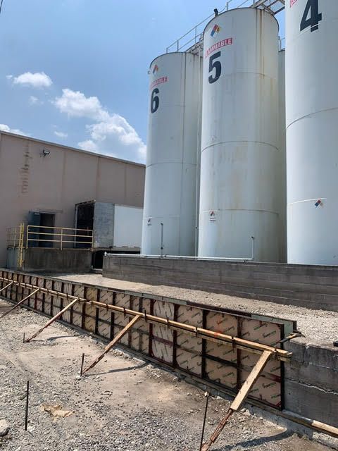 Concrete forms for a retaining wall in front of industrial storage tanks. The setting is outdoors on a cloudy day.