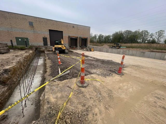 Construction site with a building, trench, heavy machinery, orange cones, and caution tape.