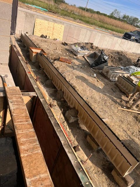 Construction site showing a long trench with wooden forms, possibly for a drainage system. Earth and building materials surround the trench.