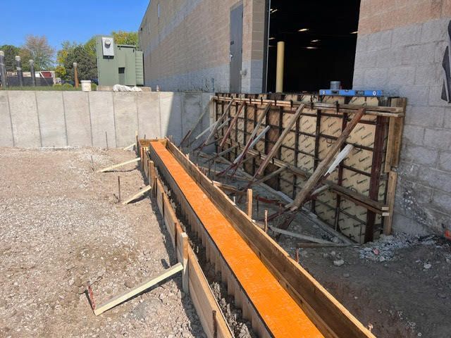 Construction site with wooden forms for concrete work. Forms run along a brick wall and are on gravel, with an open door in the background.