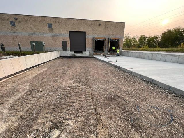 Exterior view of a loading dock with newly poured concrete, gravel driveway. Workers in high-visibility vests.