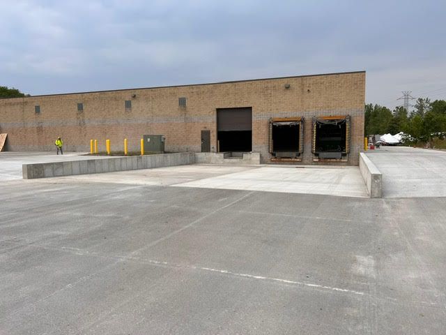 A warehouse with three loading docks; a person in a yellow vest is visible near the building. Concrete loading area and asphalt parking.