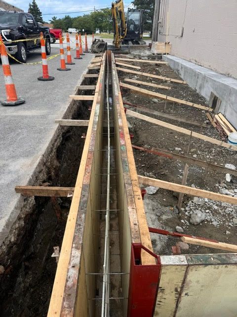 Construction of a narrow trench with wooden frames and rebar, likely for a utility line. Orange cones line the adjacent street.