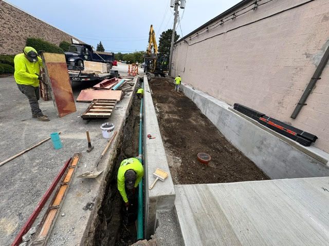 Construction workers in neon yellow vests working in a trench, next to a building and equipment.