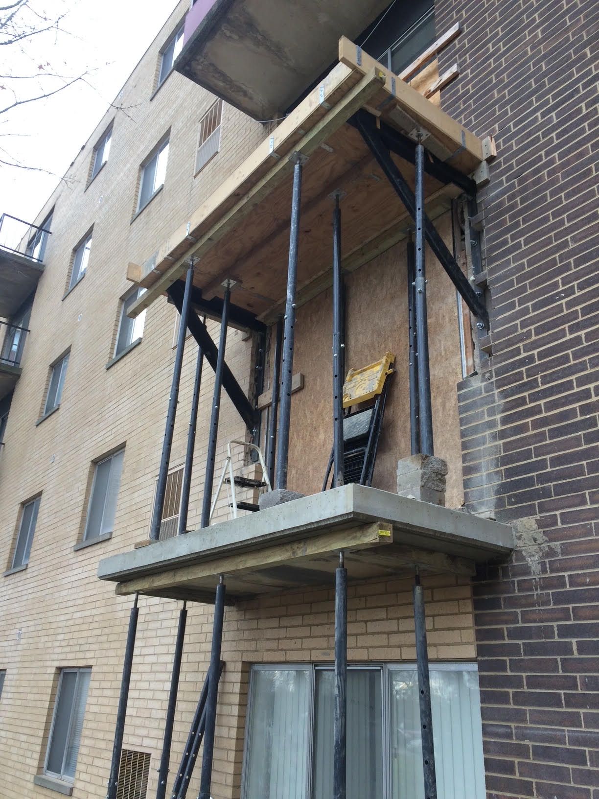 A balcony supported by scaffolding on the side of a brick building. The balcony is under repair, with a chair and supplies visible.