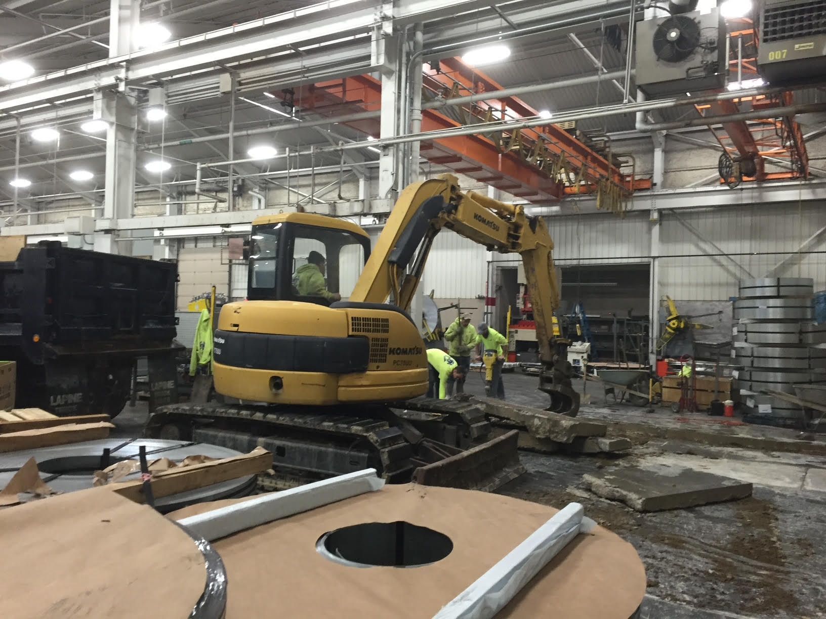 A yellow excavator dismantles concrete inside a large industrial building while workers in safety vests observe.