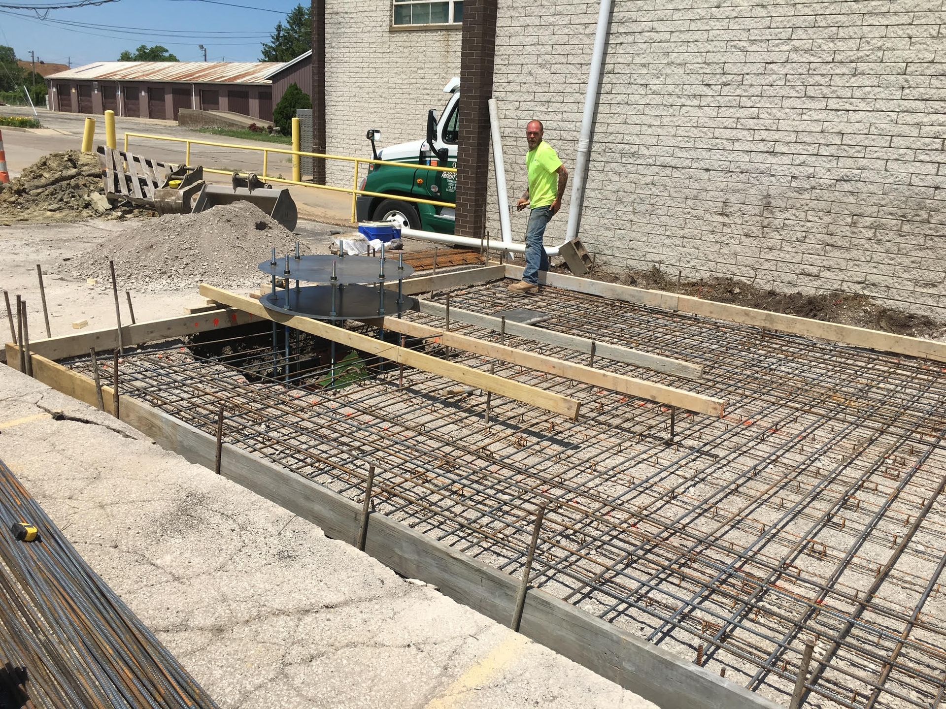 Construction site with a worker standing near a concrete form filled with rebar. Gravel and concrete forms are visible outdoors near a building.