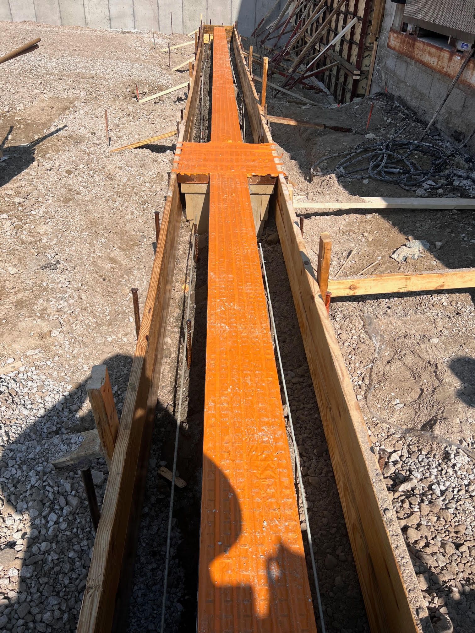 Construction site with wooden forms holding a long, narrow trench filled with orange material, possibly for a foundation. Gravel surrounds the trench.