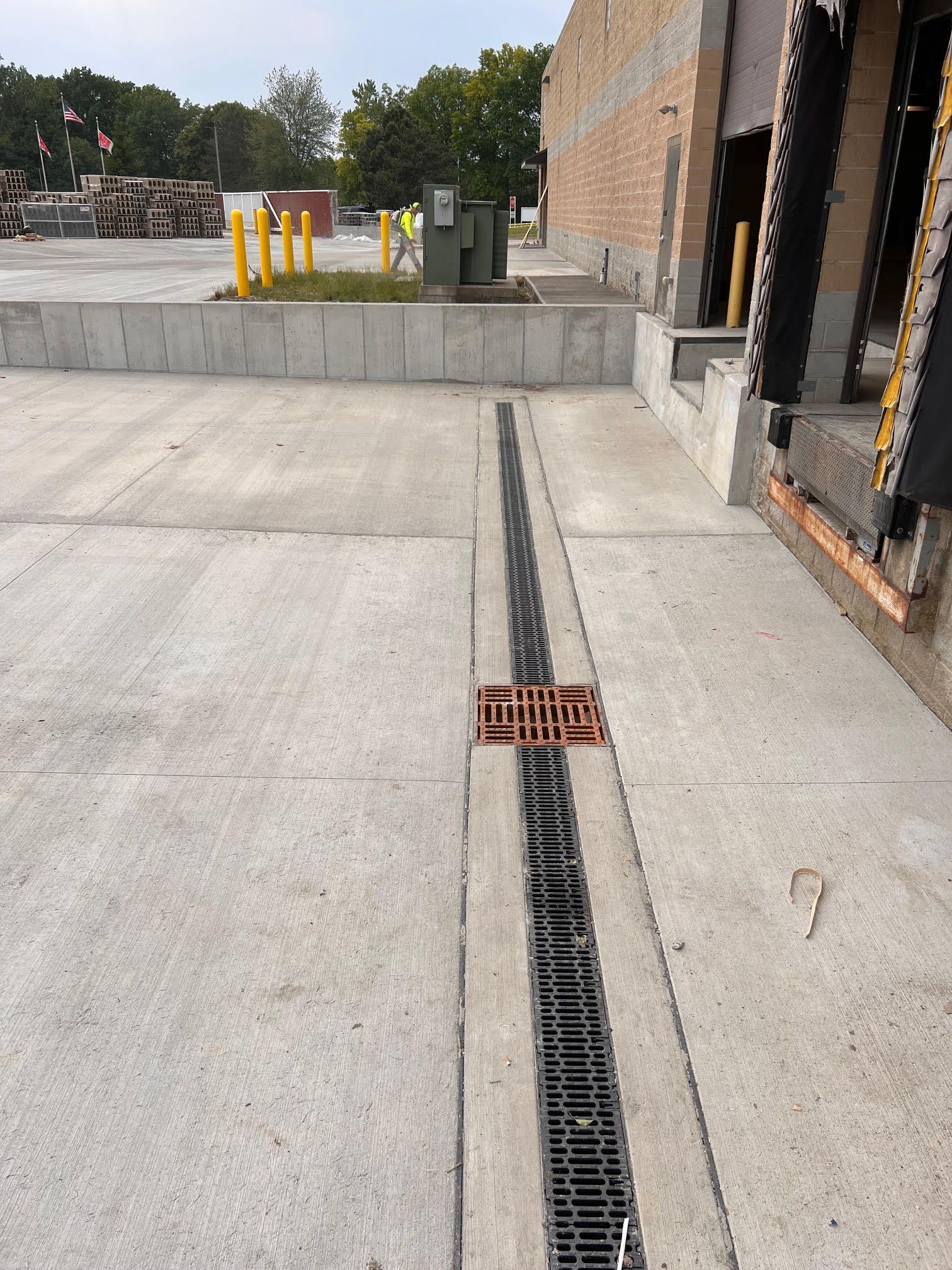 Concrete loading dock area with a long drainage grate running through it. Yellow bollards and a brick building are visible.