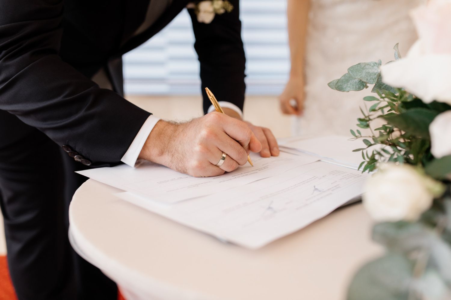 Groom in black suit signs a document at a wedding ceremony, bride in background.