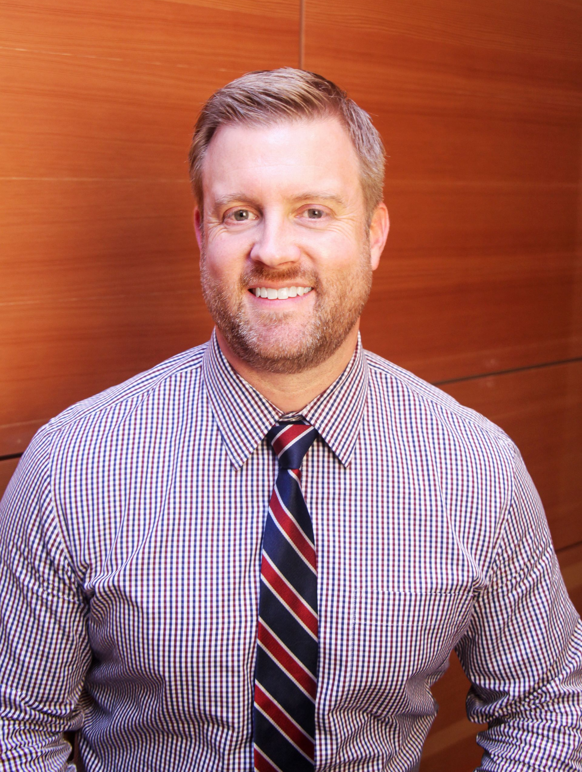 Man in a checked shirt and tie smiling against a wood-paneled wall.
