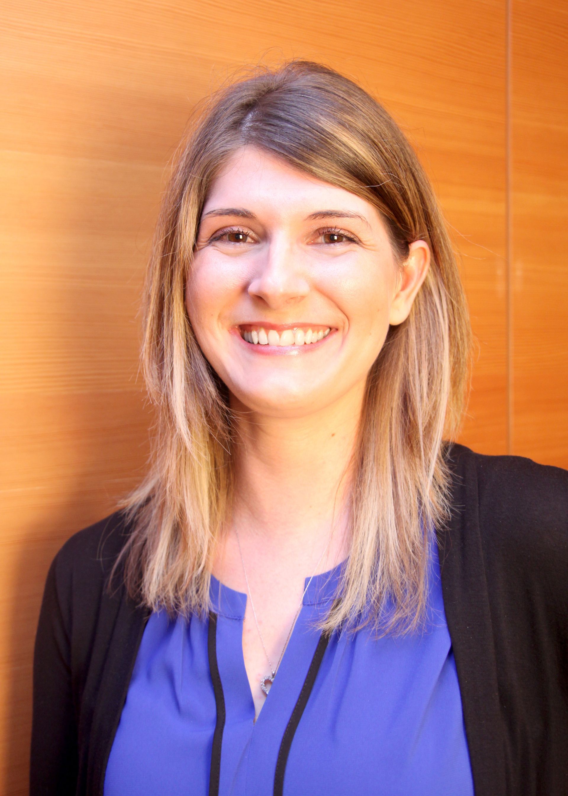 Woman with blonde hair, smiling, wearing a blue shirt and black cardigan, standing in front of a wood panel wall.