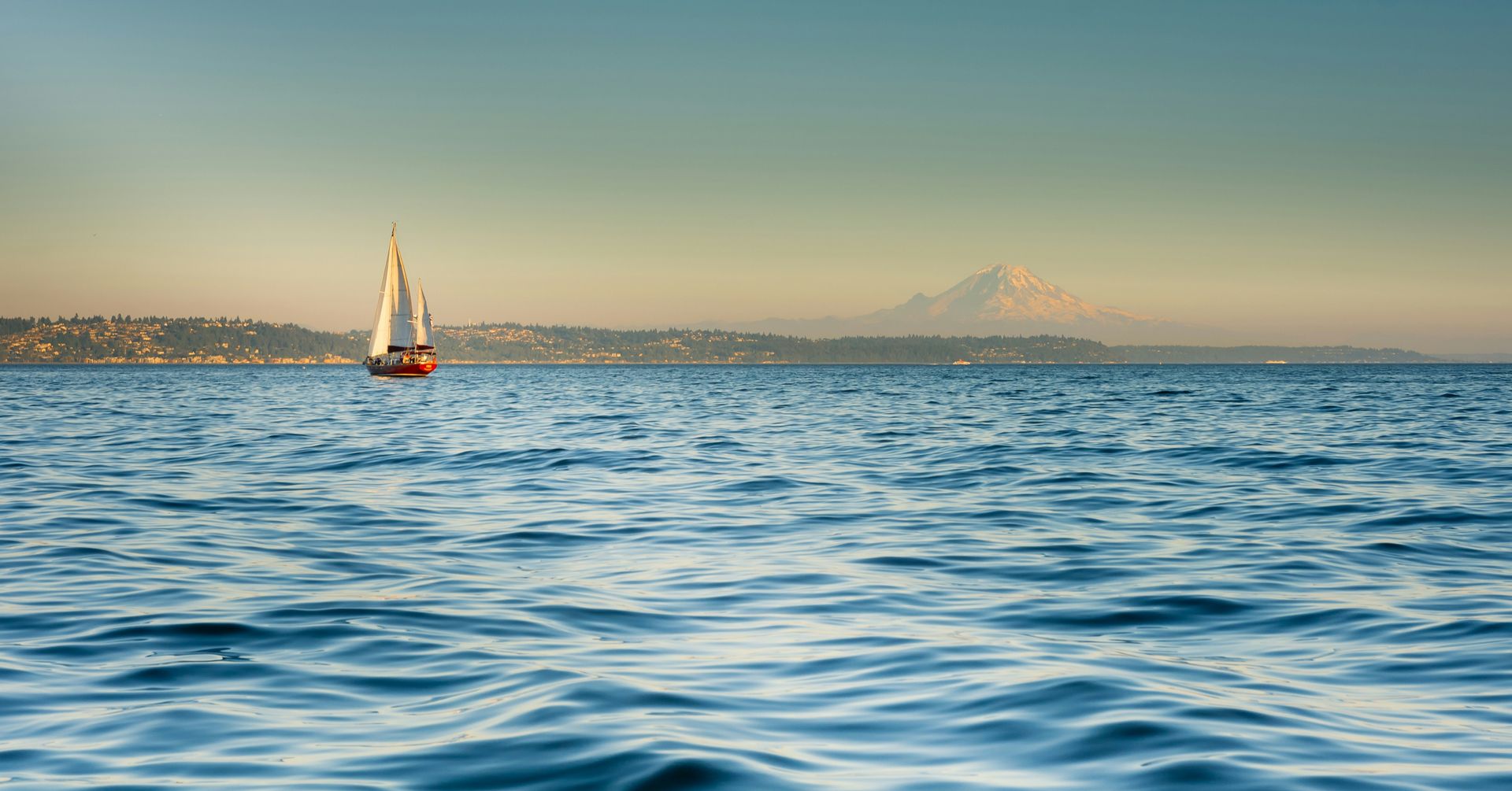 Sailboat on a wavy blue ocean under a clear, hazy sky, with a mountain range on the horizon.