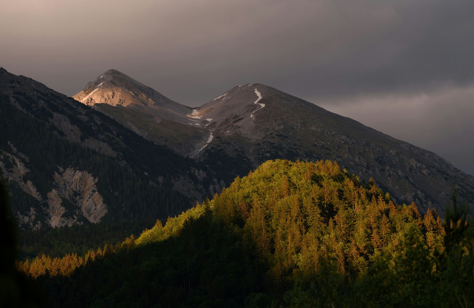 Mountains illuminated by sunlight; dark storm clouds above. Forest in the foreground.
