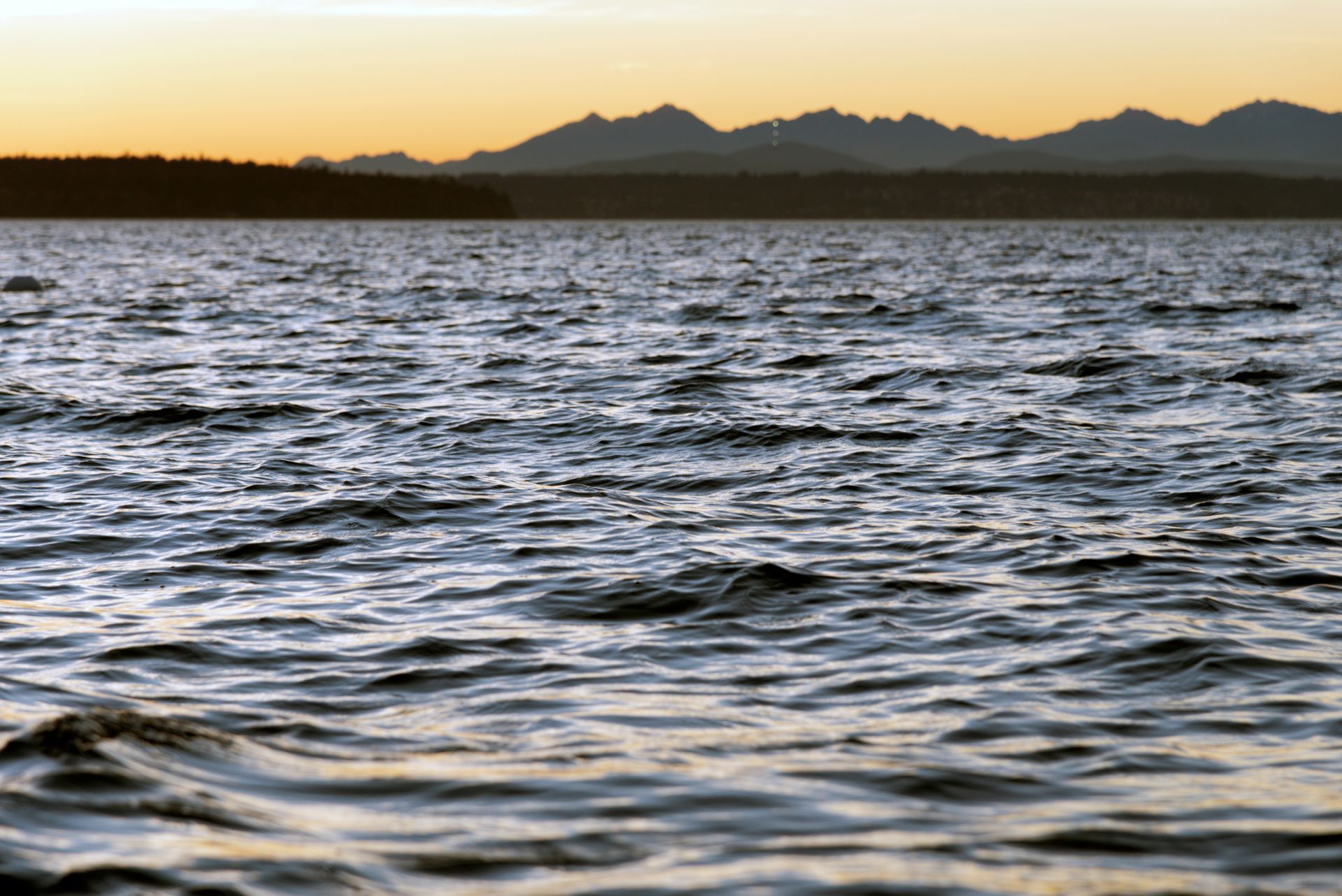 Water waves under a sunset sky, with a dark treeline and distant mountains.