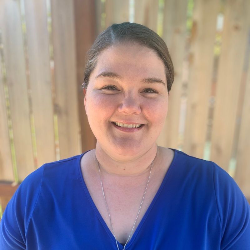 Woman with a smile in a blue shirt, standing outside in front of a wooden fence.