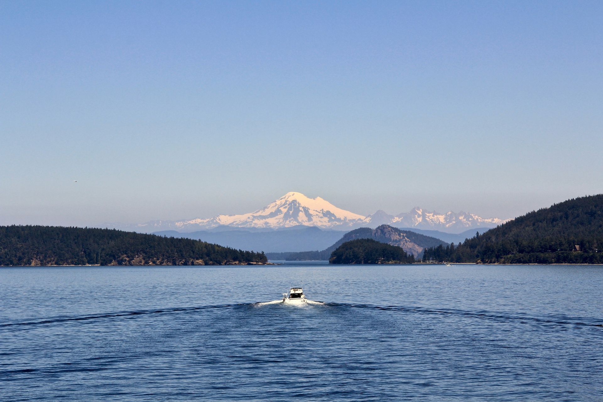 Boat on blue water with a snow-capped mountain and forested islands under a clear sky.