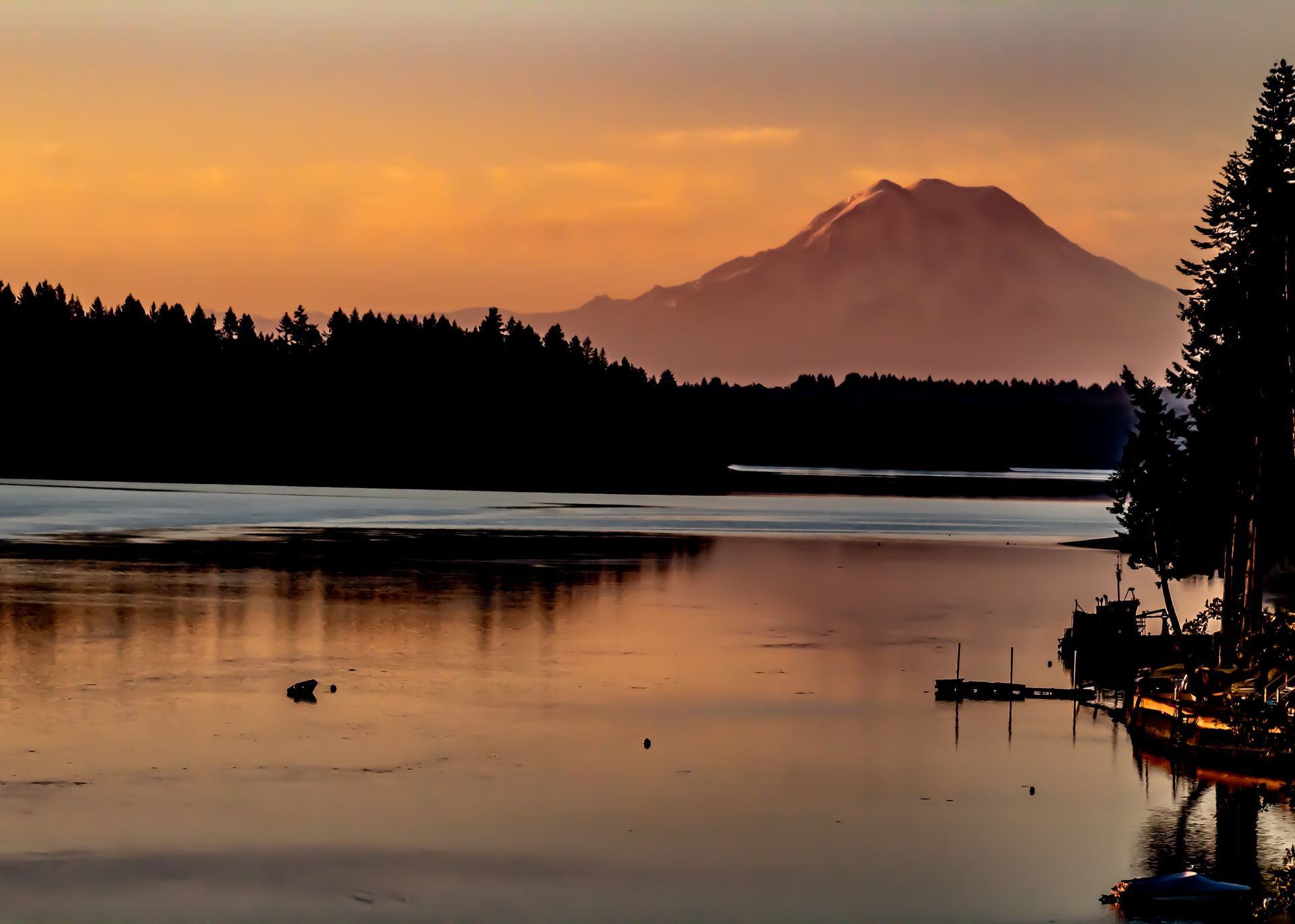 Sunset over water, silhouetted forest, mountain in distance, orange sky reflected on water.