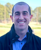 A man in a blue shirt and black jacket is smiling for the camera.