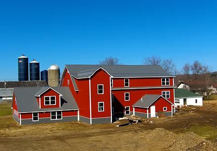 Red farm buildings with gray trim and roofs against a blue sky, silos in the distance.