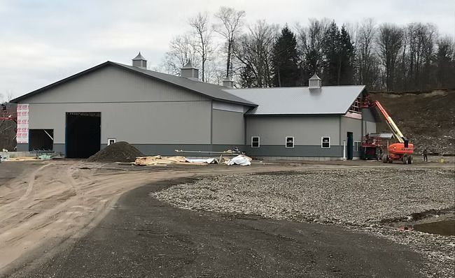 A large gray metal building under construction, with an orange lift. Gravel driveway.