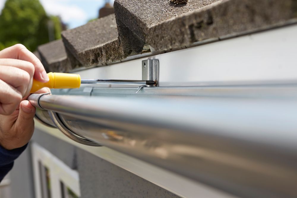 Person using a yellow screwdriver to attach a metal gutter to a house, close-up.