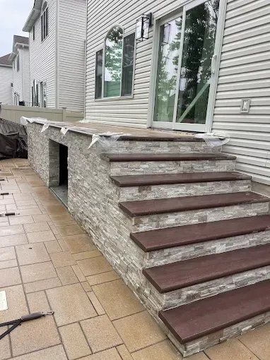 Stone steps lead up to a raised patio with a modern gray stone facade. Brown composite steps.