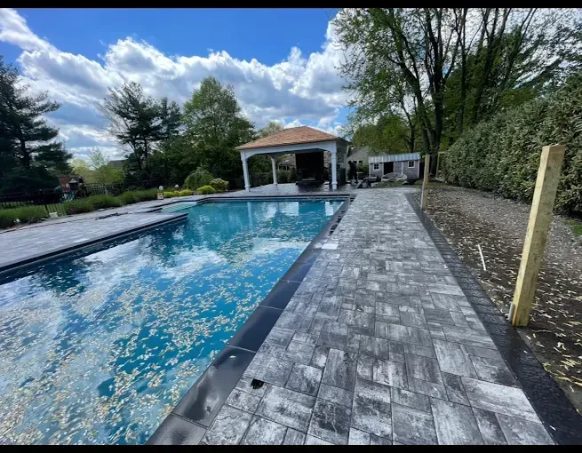 Pool with brick walkway, gazebo, and surrounding greenery under a blue sky.