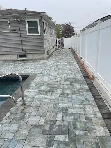 A gray-paved path alongside a pool and white fence, leading towards a gate between two buildings.