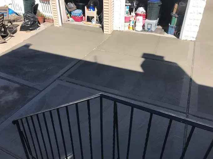 New concrete driveway and walkway with a black railing in the foreground; garage door open.