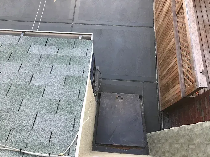 Gray wall with a green shingle roof, a black metal cover, and a wooden latticed structure.