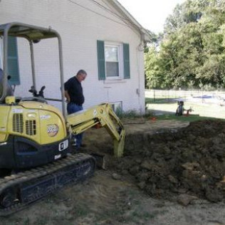 A man is standing next to a yellow excavator in front of a house.