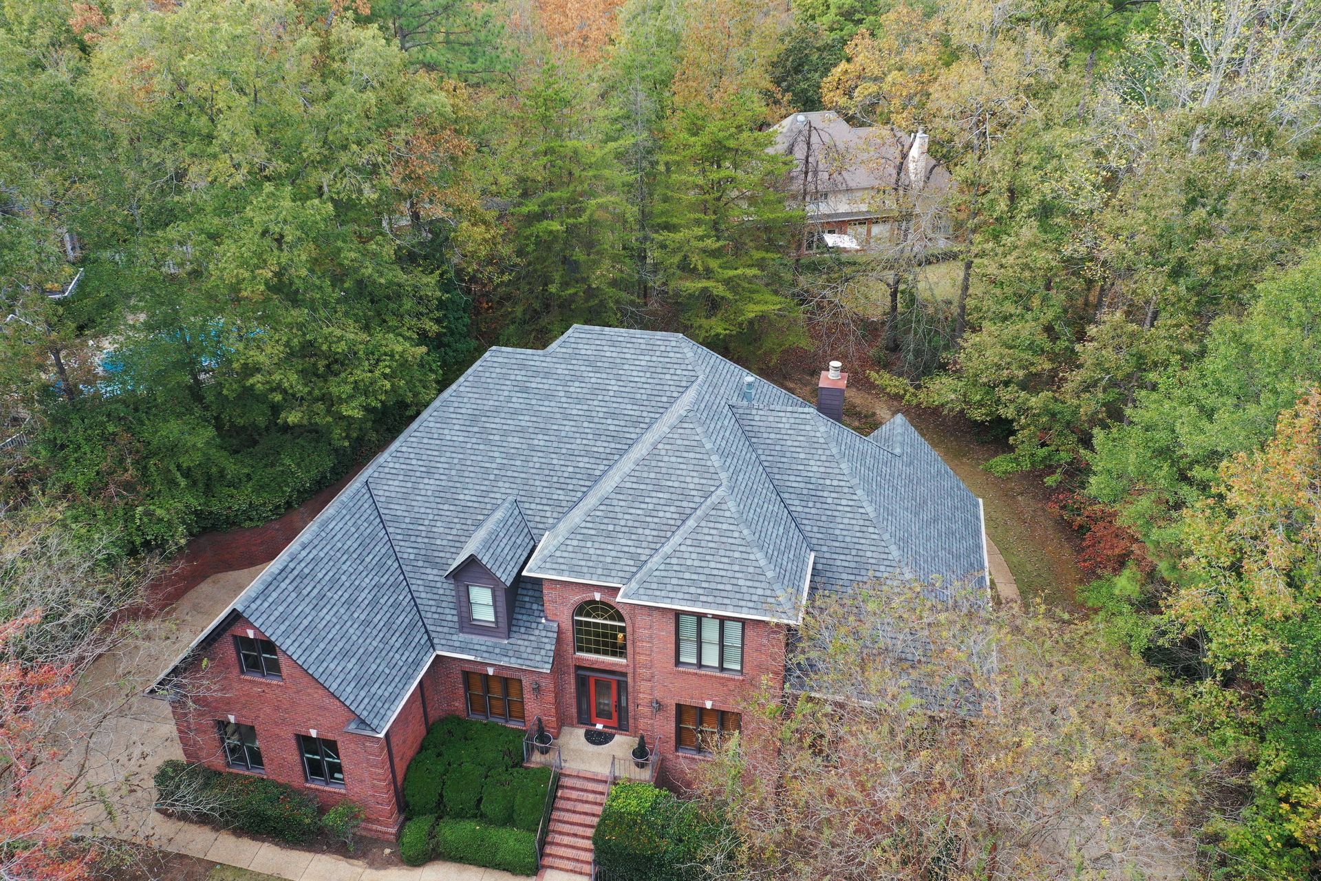 An aerial view of a red brick house with a gray shingled roof, surrounded by trees during autumn.