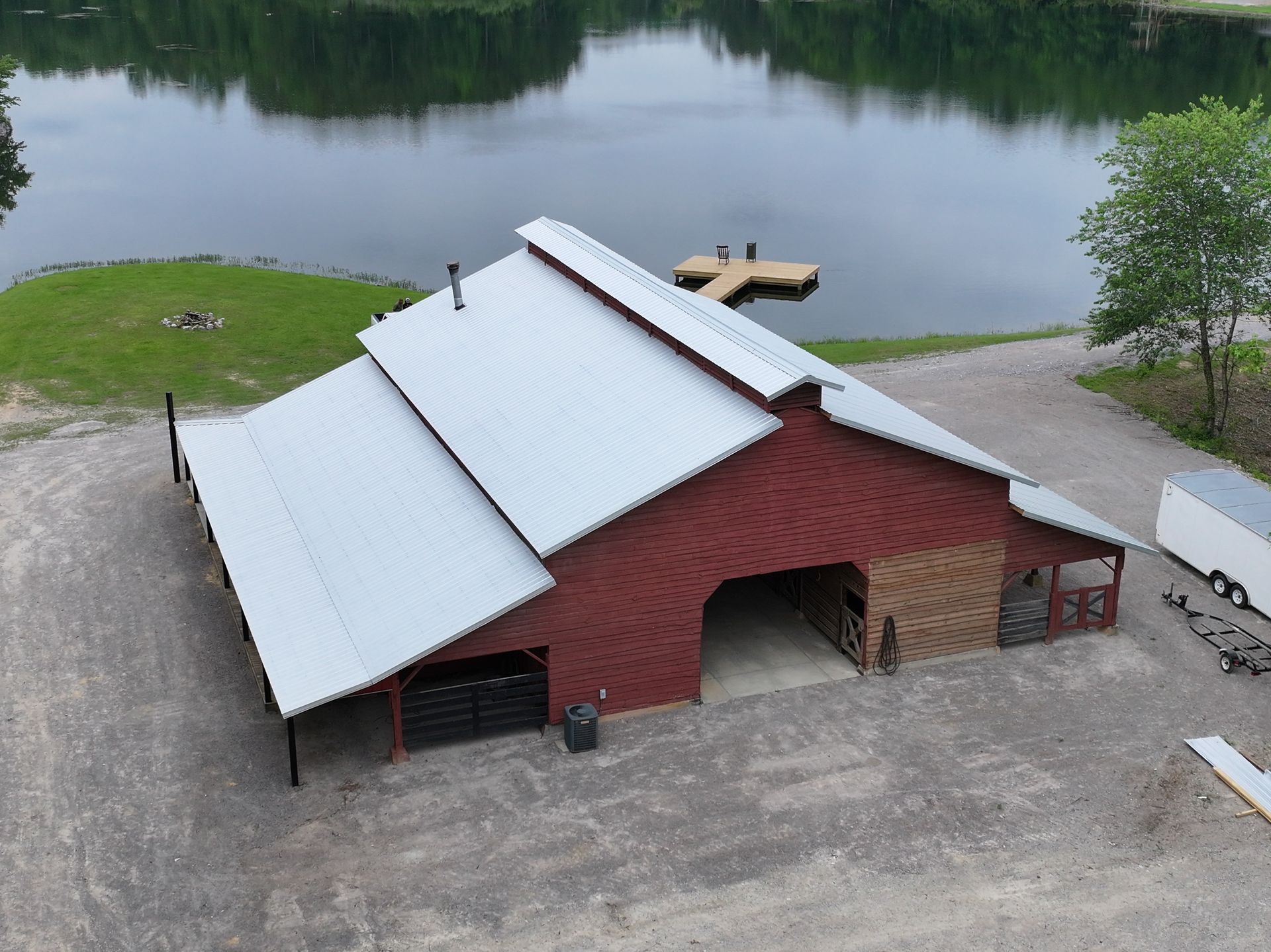 An aerial view of a red barn with a metal roof, situated on a gravel lot next to a lake with a wooden dock.