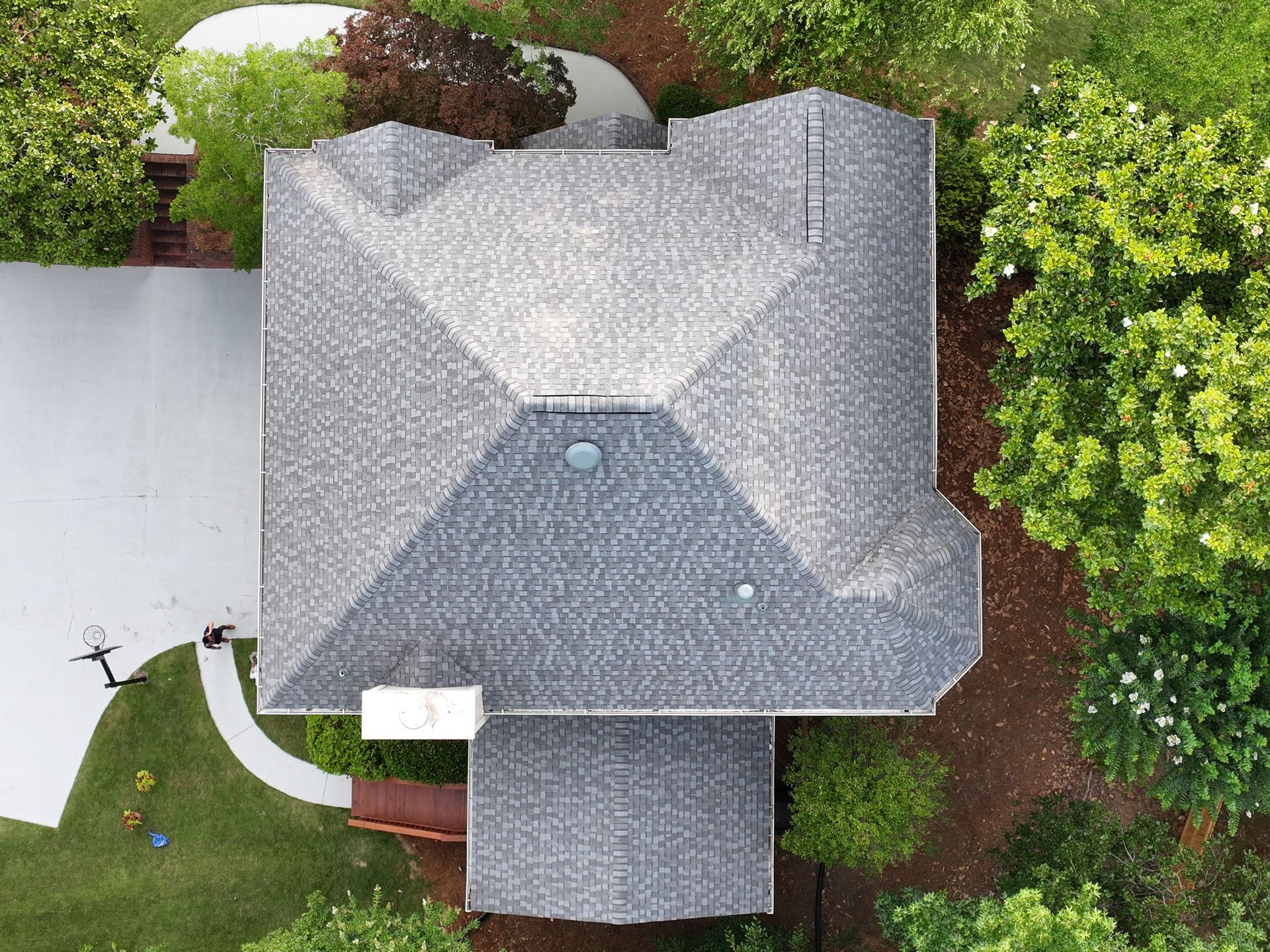 Top-down aerial view of a gray shingled residential roof surrounded by green trees and a driveway.