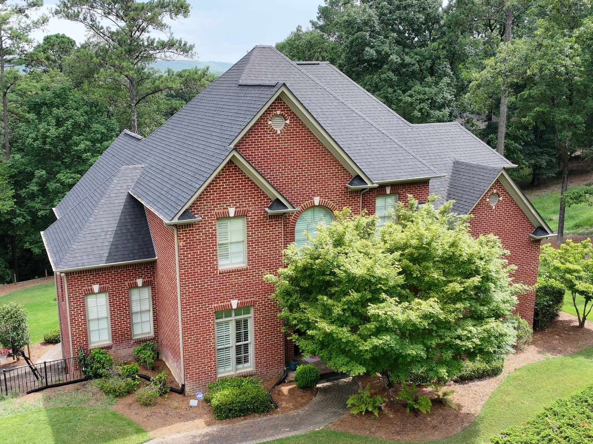 An aerial view of a two-story red brick house with a gray shingle roof, surrounded by green trees and a lawn.