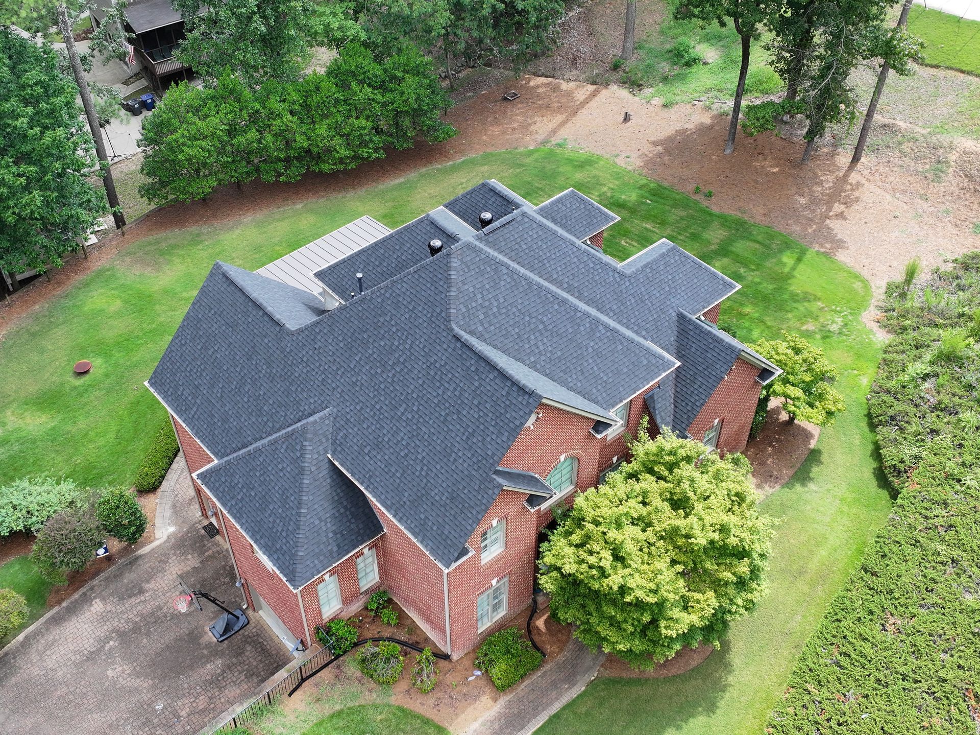 A high-angle aerial view of a multi-gabled red brick house with a dark grey shingled roof, surrounded by a grassy lawn.
